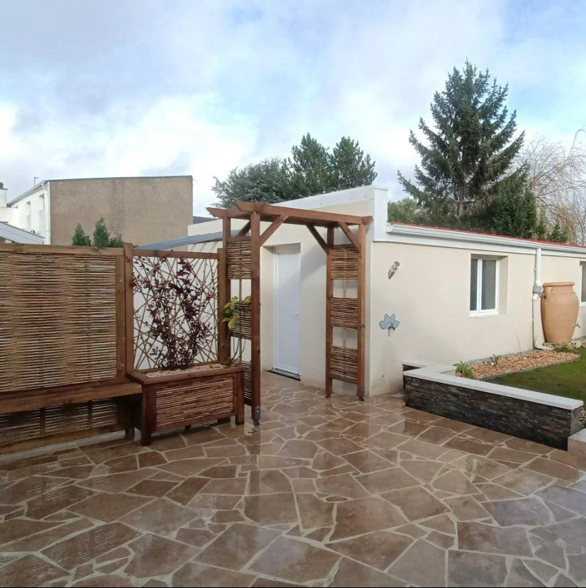 Arche de jardin en bambou et bois avec structure carrée et lucarnes, situé sur une terrasse pavée, près d’un mur blanc et d’un grand jarre décoratif. Fabrication artisanale française.